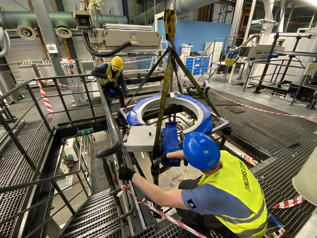 Dwóch serwisantów w kaskach i kamizelkach, jeden z widocznym logo 'TINCORS.COM', instaluje niebieski element maszyny na platformie z metalowej kraty, używając dźwigu w hali przemysłowej.