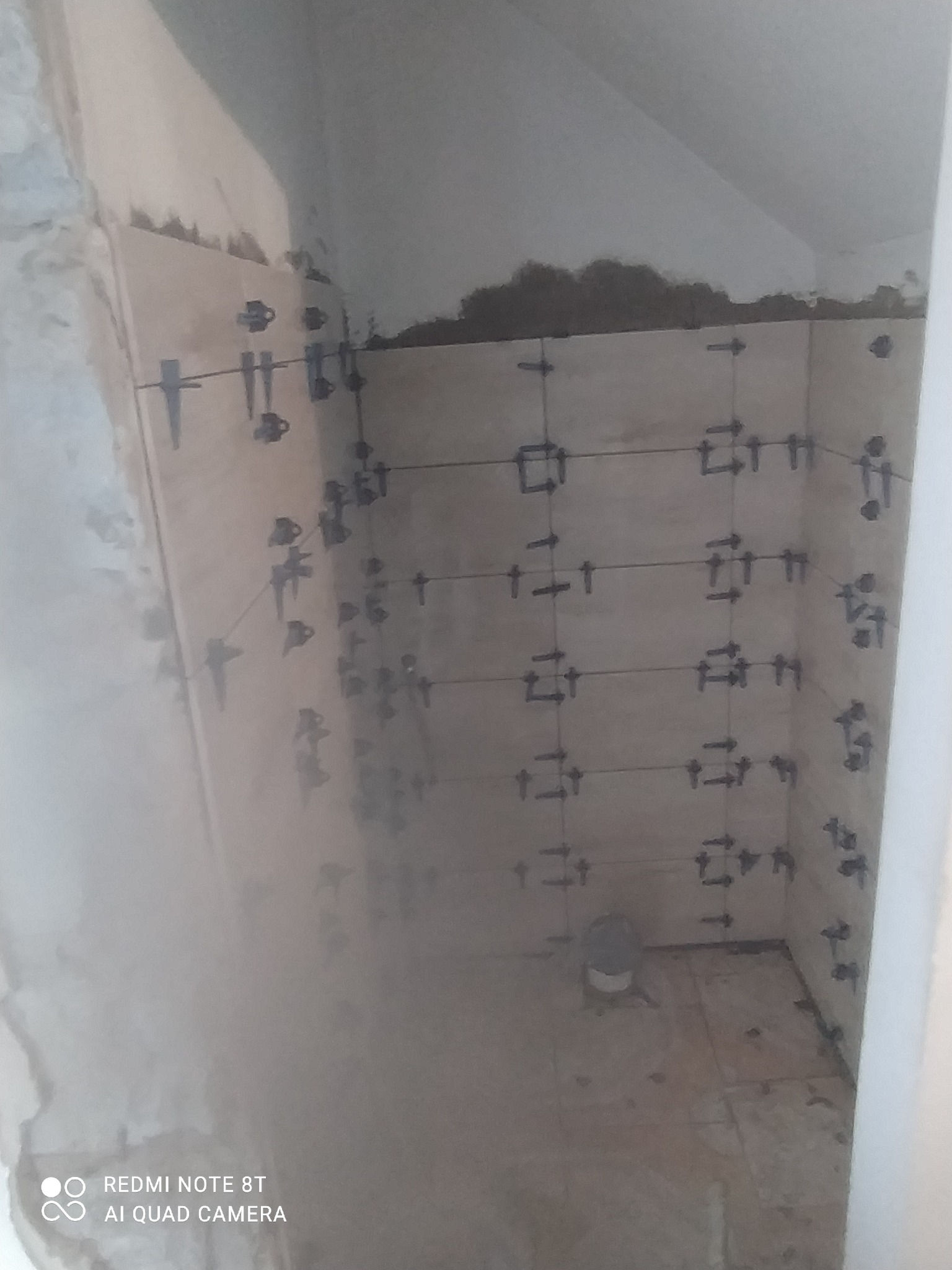 Świeżo ułożone płytki ścienne z krzyżykami dystansowymi w trakcie remontu łazienki, widok na narożnik i fragment podłogi z pozostałościami zaprawy.