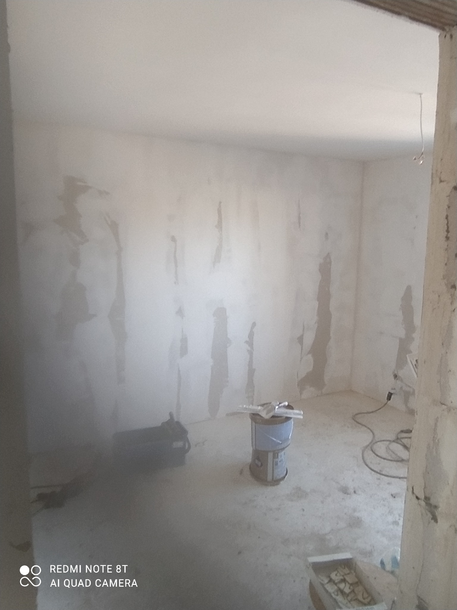 Pokój w trakcie prac wykończeniowych, ściany z widocznymi śladami szpachlowania, narzędzia malarskie i wiadro z farbą na podłodze, widoczny kabel elektryczny.