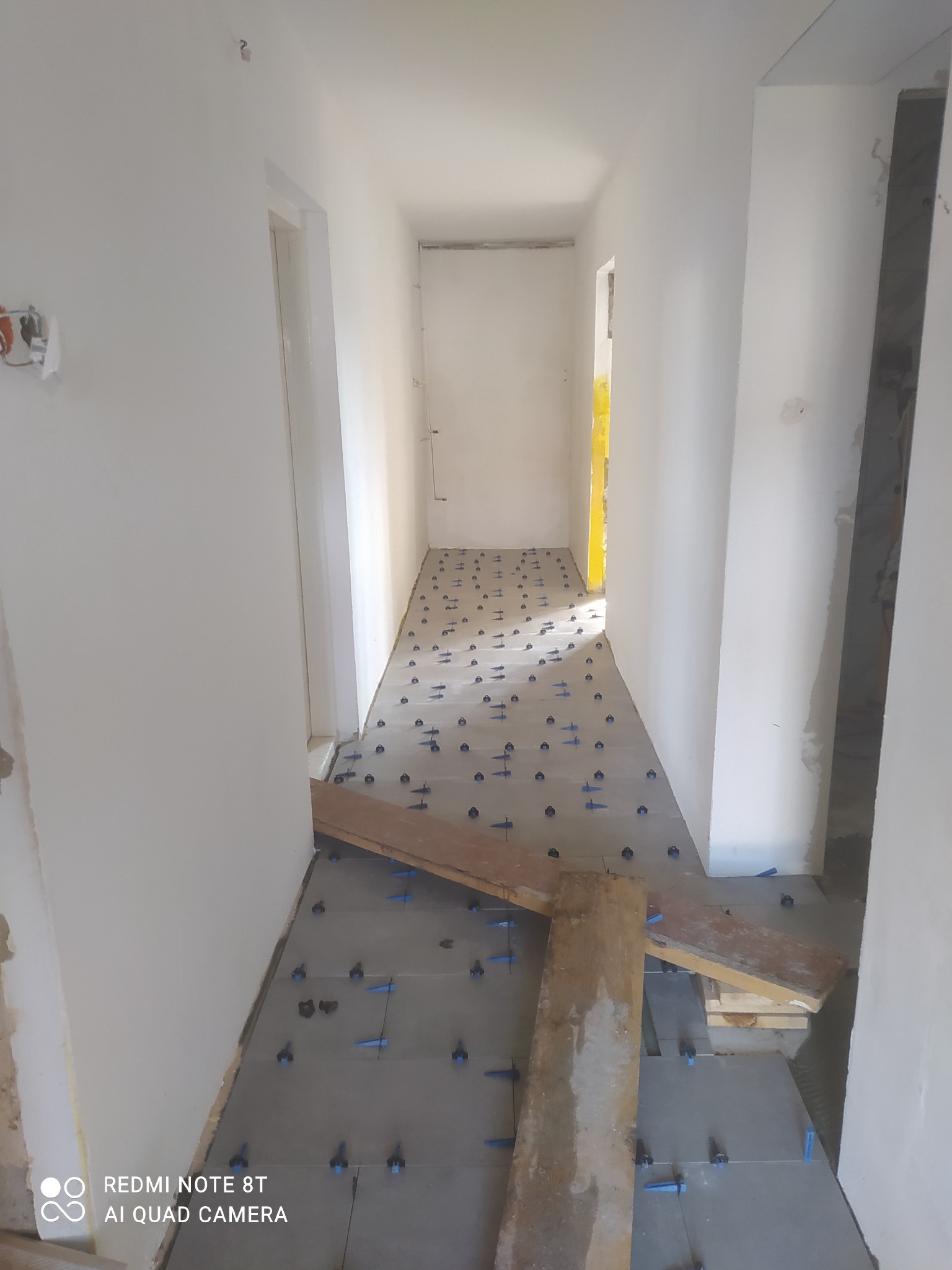 Korytarz w trakcie układania szarych płytek podłogowych z zastosowaniem niebieskich krzyżyków dystansowych i drewnianych desek jako pomostu.