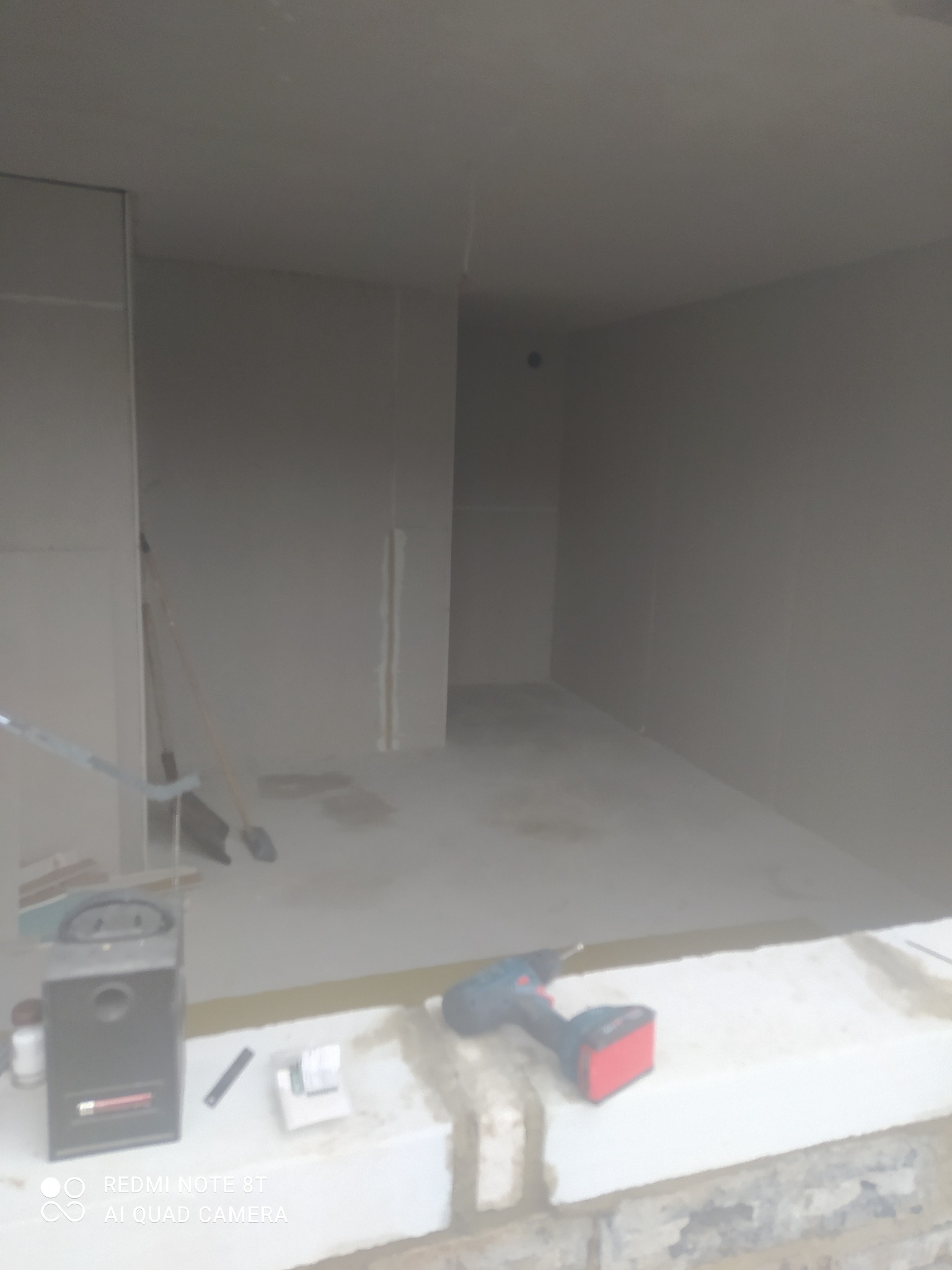 Surowe wnętrze w trakcie wykończenia, ściany pokryte szpachlą, widoczna wiertarka akumulatorowa i narzędzia na parapecie, niedoskonałości i zapylenie podkreślają etap prac.