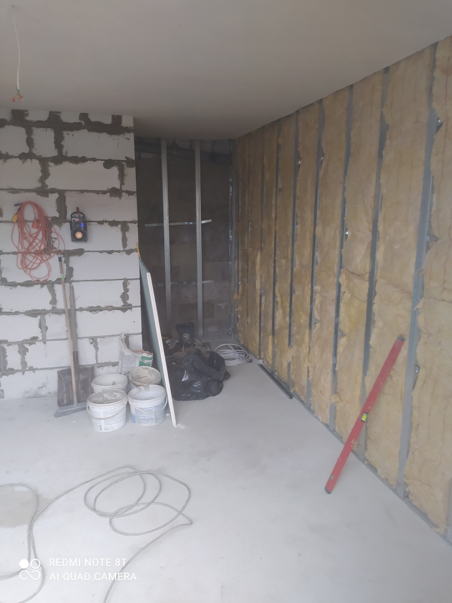 Wnętrze w trakcie prac remontowych: ściana z izolacją z wełny mineralnej w metalowej konstrukcji, surowa ściana z bloczków betonowych, wiadro farby, poziomica i zwój kabla na podłodze.