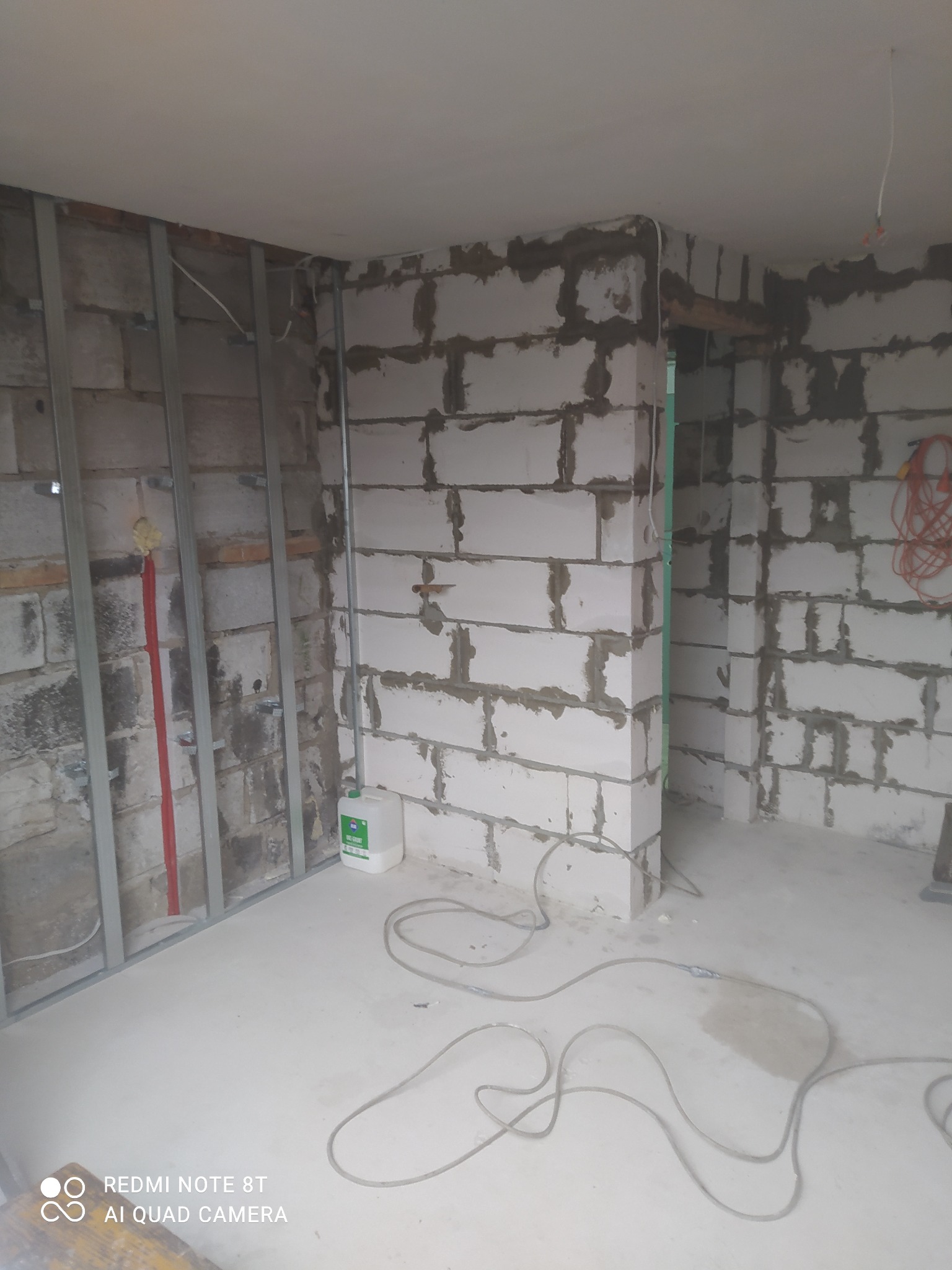 Wnętrze w trakcie prac wykończeniowych: ściany z białych bloczków z widocznymi spoinami, metalowy stelaż do zabudowy, instalacje elektryczne, biała podłoga z kablami i pojemnik z płynem...