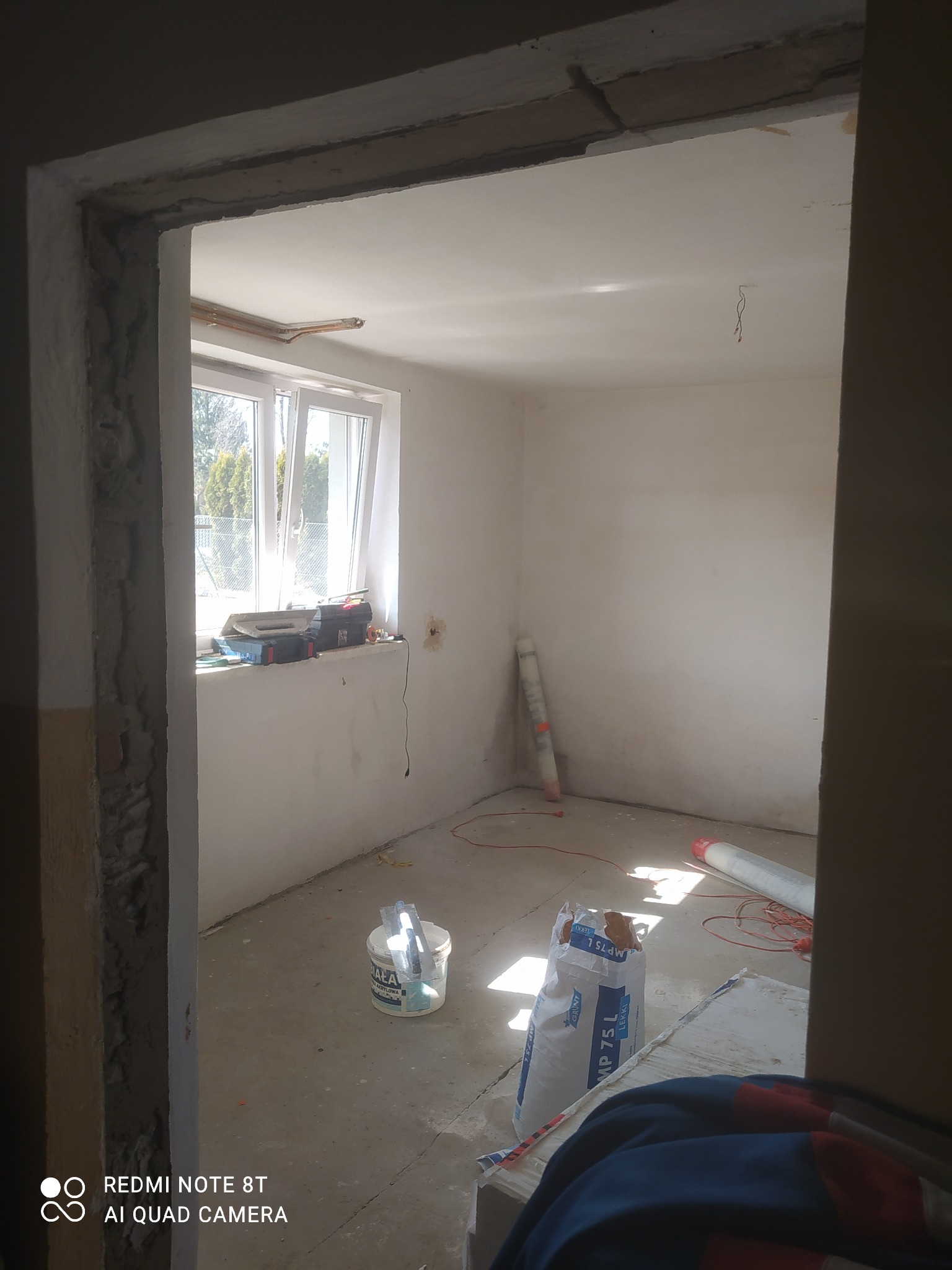 Wnętrze pokoju w trakcie remontu: surowe ściany, okno z narzędziami, wiadro z zaprawą, worek gipsu MP 75 L oraz przewody elektryczne na podłodze.