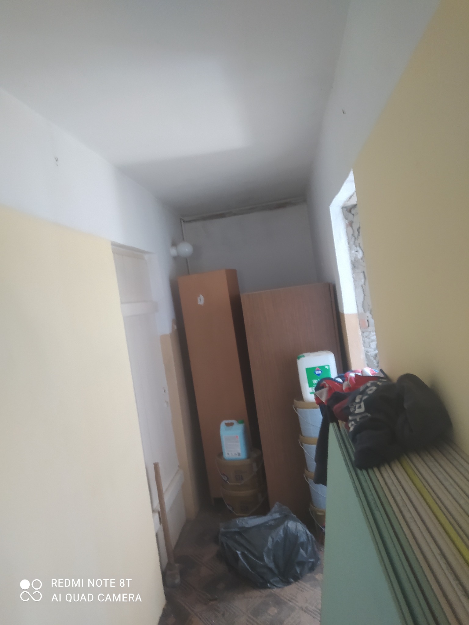 Korytarz w trakcie remontu: widoczne płyty gipsowo-kartonowe, farby, narzędzia i worki na śmieci. Fragment ściany z odsłoniętym murem przy oknie.