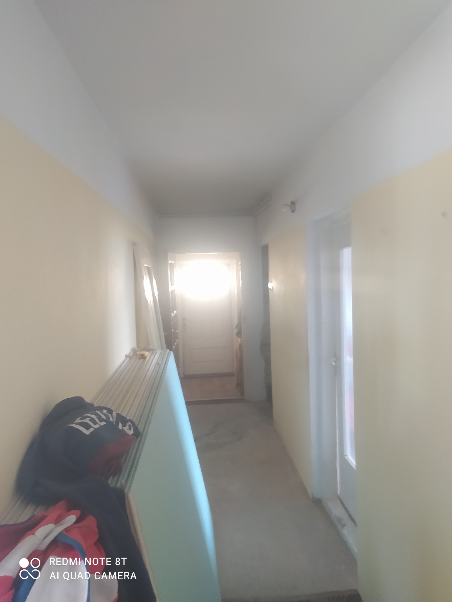 Korytarz w trakcie remontu: ściany przygotowane do malowania, widoczne płyty gipsowe oparte o ścianę, ubrania robocze na wierzchu, drzwi wewnętrzne po prawej stronie.