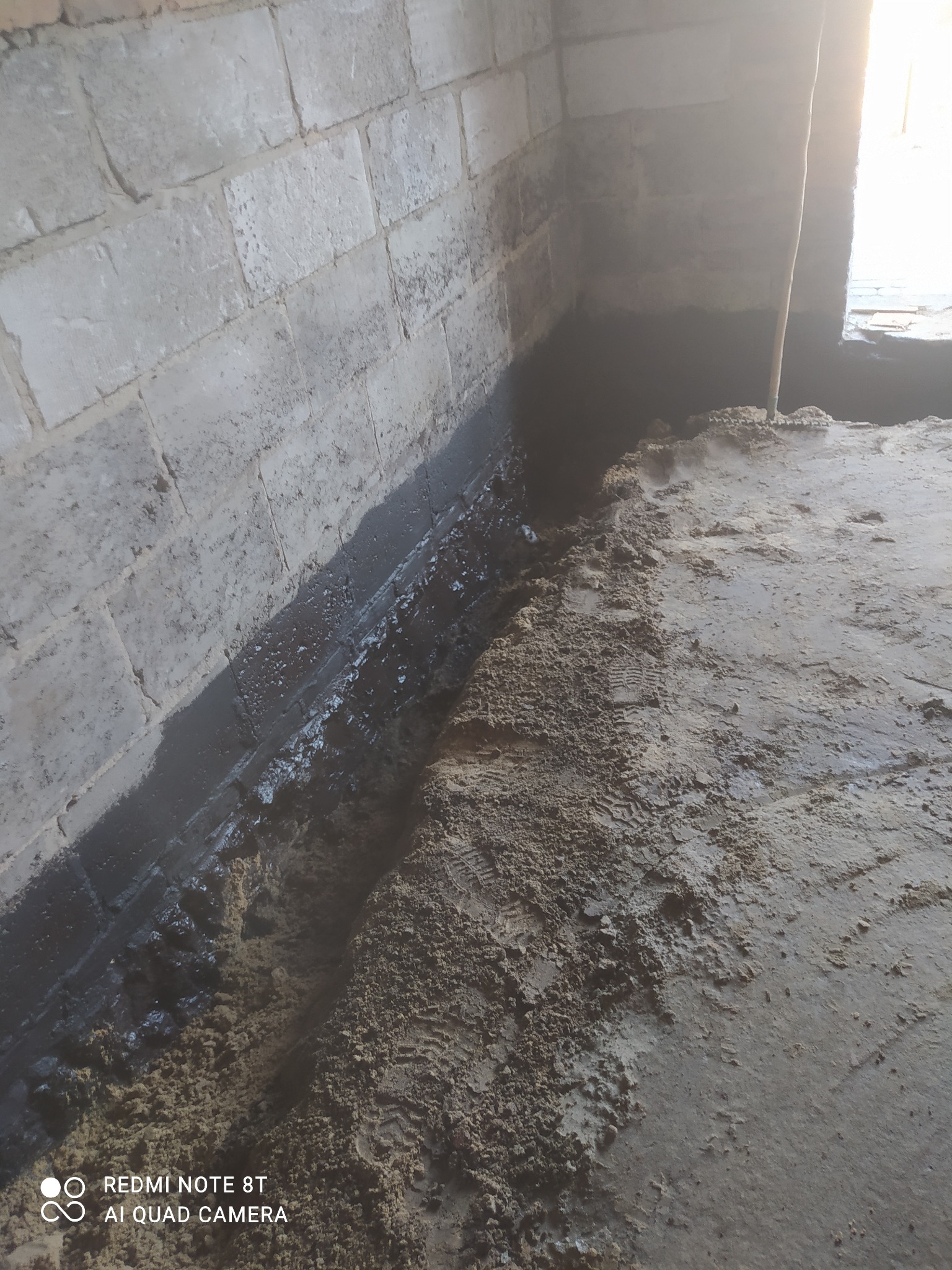 Hydroizolacja fundamentów budynku z bloczków betonowych, widoczna warstwa bitumiczna na styku ściany i posadzki, podłoże z piasku i ślady obuwia.