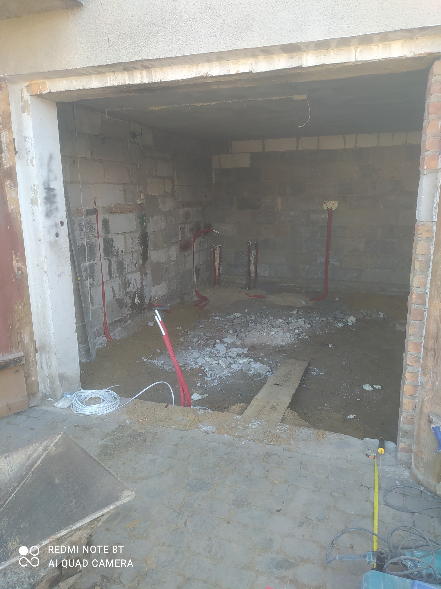 Wnętrze remontowanego pomieszczenia z odsłoniętymi przewodami instalacji elektrycznej i wodnej, gruzem na podłodze i narzędziami budowlanymi w tle.