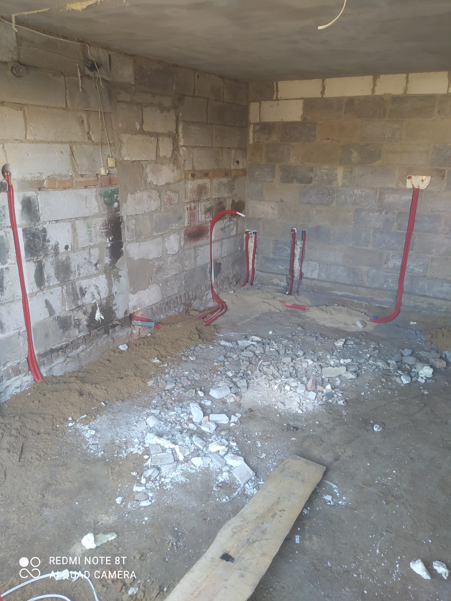 Surowy stan wnętrza w trakcie prac instalacyjnych: ściany z bloków, czerwone rury instalacji wodnej wystające ze ścian, gruz i piasek na podłodze, widoczny fragment deski.