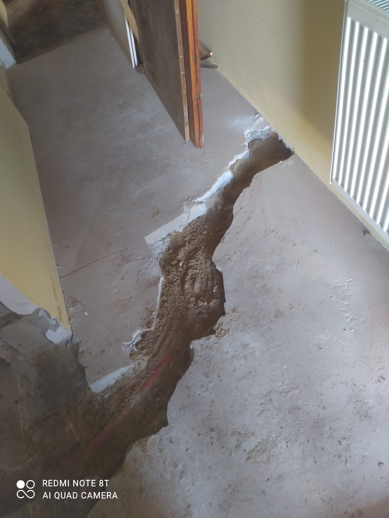 Wykop w posadzce betonowej z widocznymi rurami instalacji CO, ślady gruzu i pyłu budowlanego, grzejnik żeberkowy w tle.