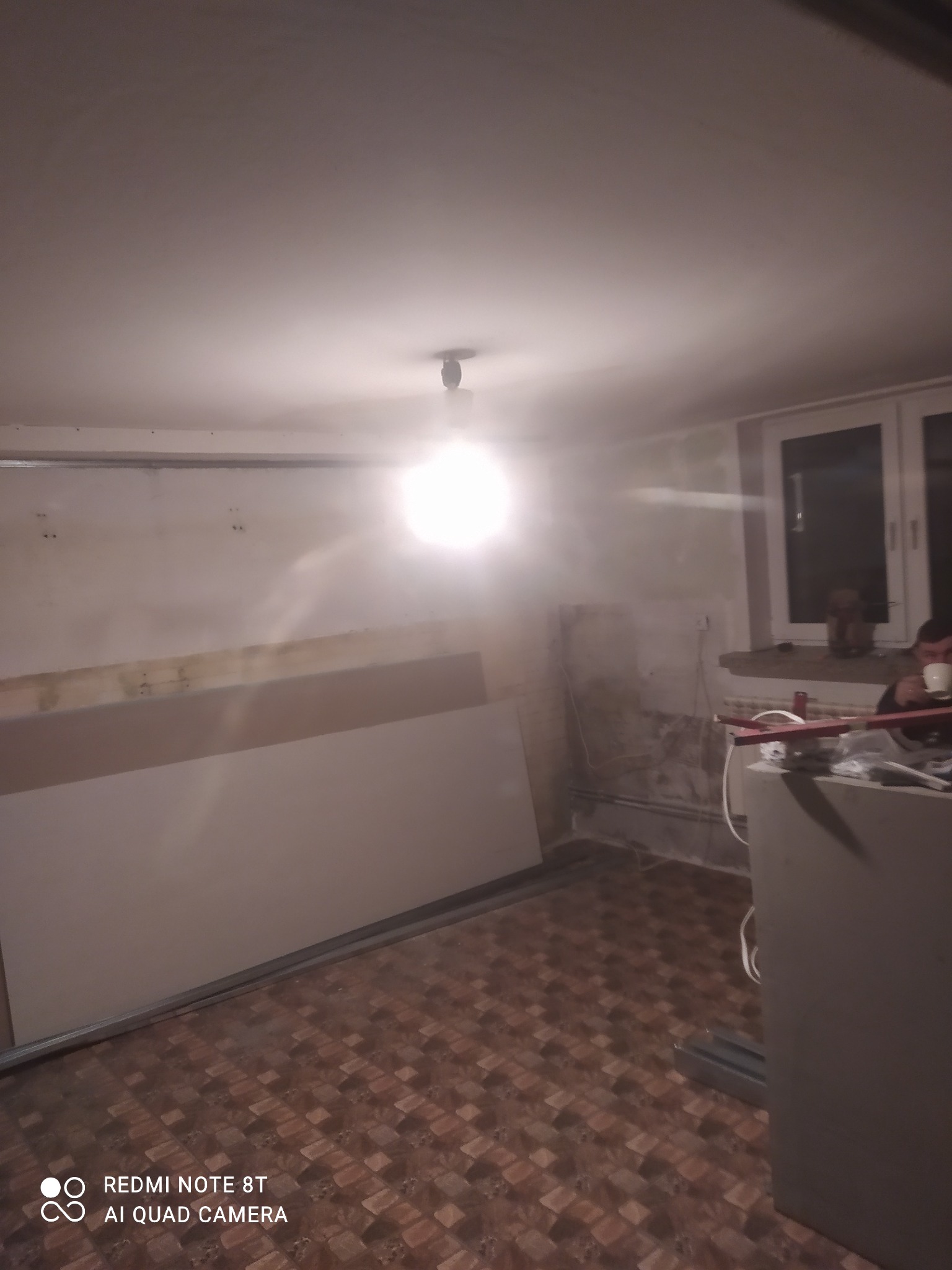 Wnętrze pokoju w trakcie remontu: płyty gipsowe oparte o ścianę, widoczne ślady po wcześniejszych pracach, mężczyzna pije kawę w tle przy oknie.