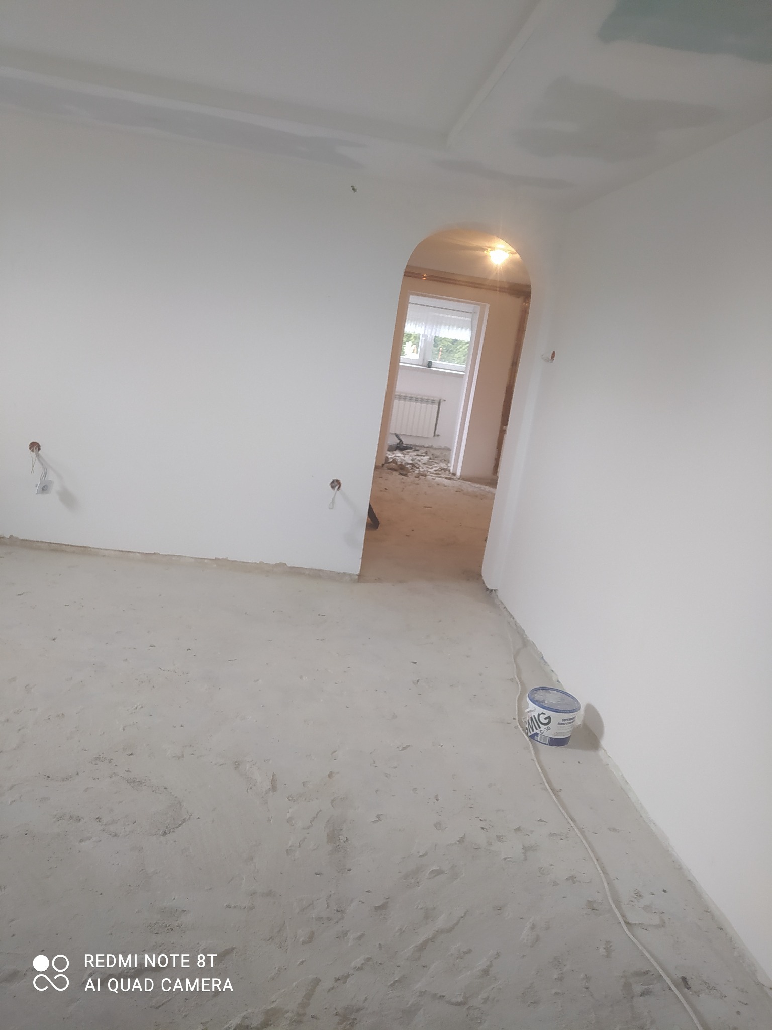 Wnętrze w trakcie remontu: surowe ściany, podłoga z resztkami zaprawy, widoczne puszki elektryczne bez osprzętu, przejście łukowe do kolejnego pomieszczenia z gruzem i grzejnikiem, wiadro farby...