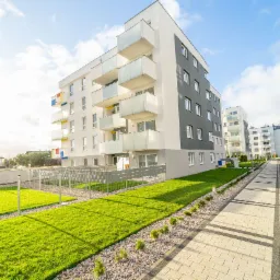 Nowoczesny kompleks apartamentowy z balkonami, utrzymany w bieli i szarości, z zadbanym trawnikiem i chodnikiem z kostki brukowej.