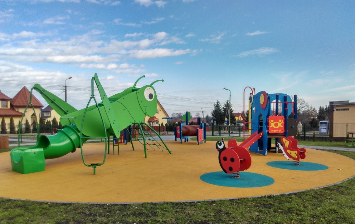 Nowoczesny plac zabaw z zielonym konikiem polnym ze zjeżdżalnią, czerwonymi biedronkami na sprężynach i niebiesko-czerwonym zestawem zabawowym na żółtej nawierzchni, na tle domów i błękitnego nieba.