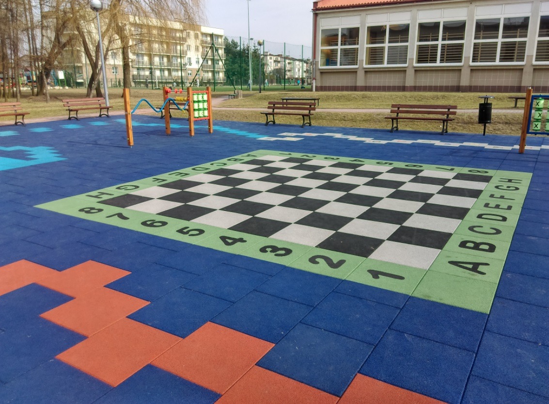 Szachownica narysowana na gumowej nawierzchni placu zabaw z literami i cyframi na obrzeżach, w tle budynek i drzewa.