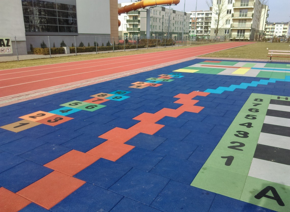 Nawierzchnia placu zabaw z gumowych płyt w kolorze niebieskim, z namalowanymi grami: klasy, zygzak z czerwonych kwadratów, szachownica. W tle bieżnia i budynki.