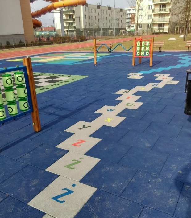Niebieska nawierzchnia placu zabaw z edukacyjną ścieżką literową i elementami do gry w kółko i krzyżyk oraz liczydło walcowe, w tle budynki mieszkalne.