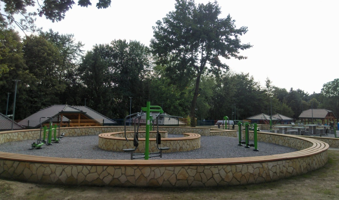 Ogródek jordanowski z zielonymi urządzeniami do ćwiczeń, otoczony kamiennym murkiem z drewnianym siedziskiem, na żwirowej nawierzchni, z widocznymi altanami w tle.