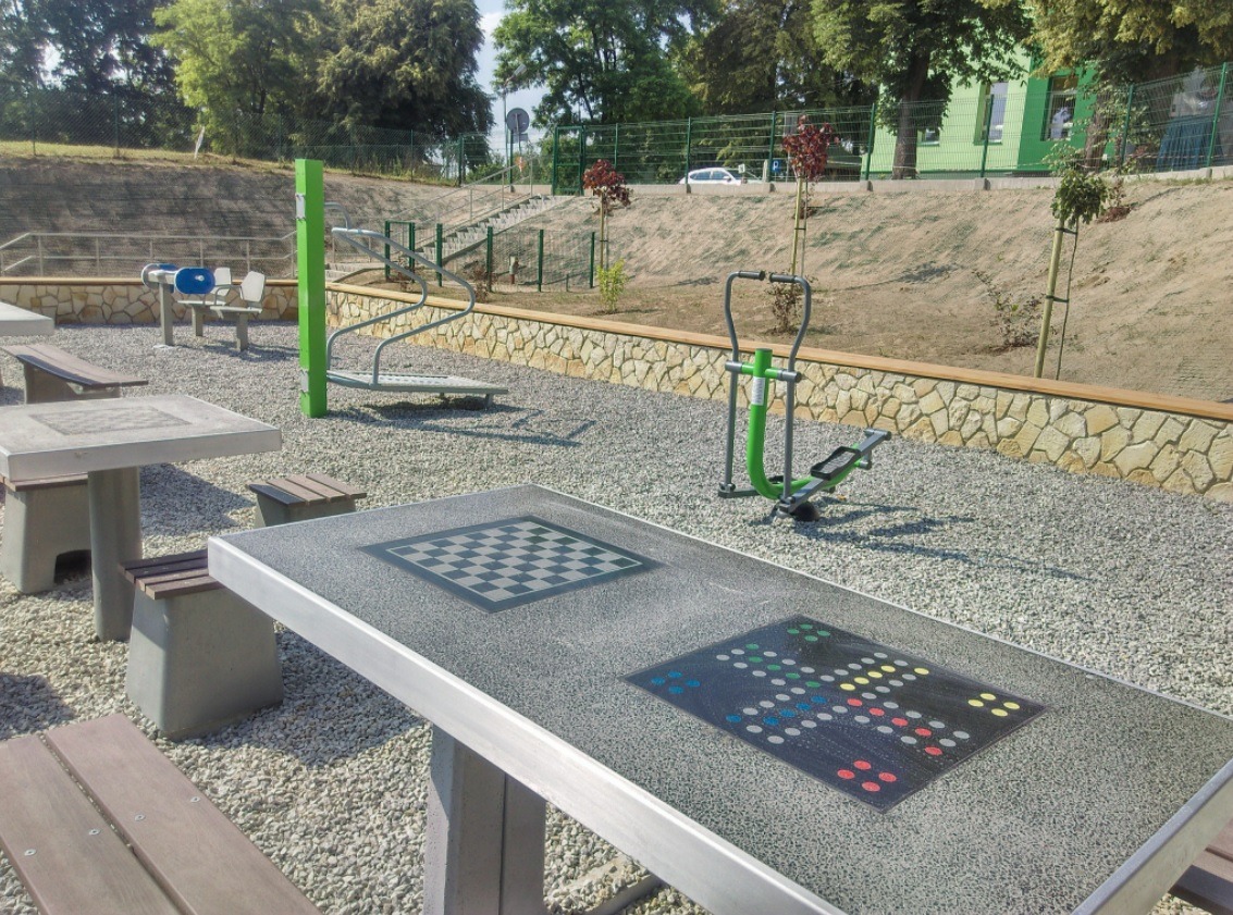 Stół do gier plenerowych z szachownicą i planszą do chińczyka, na żwirowym podłożu placu zabaw, w tle urządzenia do ćwiczeń i schody.