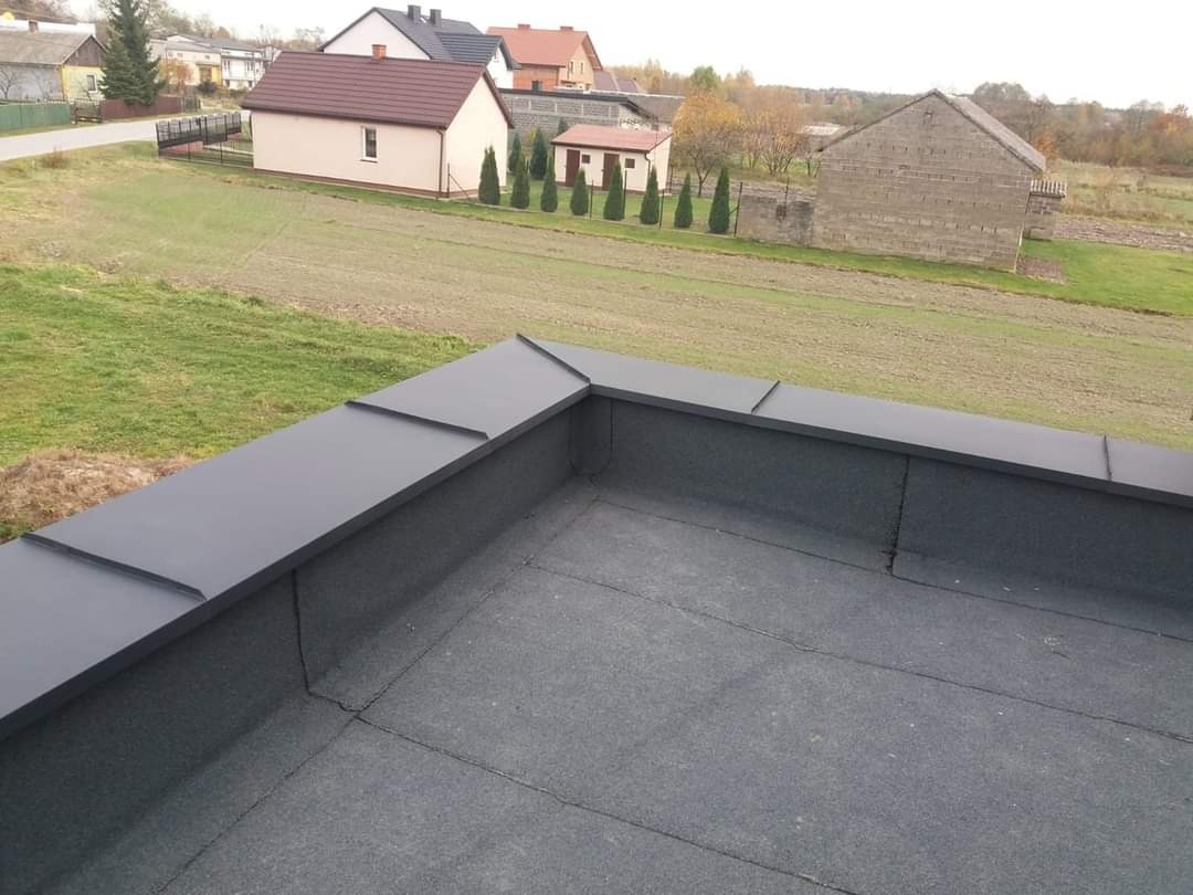 Widok z góry na płaski dach pokryty papą, z obróbkami blacharskimi w kolorze grafitowym, na tle wiejskiego krajobrazu z domami.