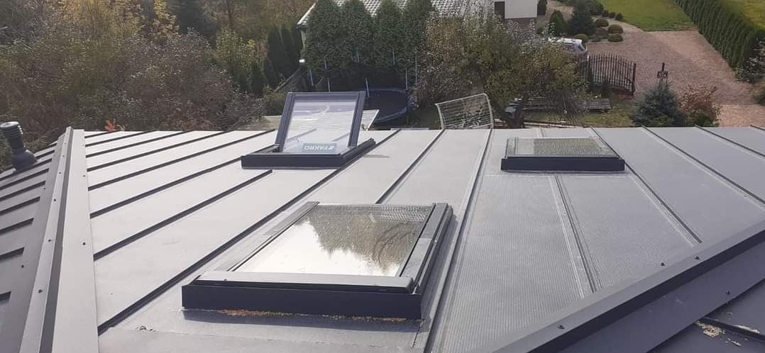 Szary dach z blachy trapezowej z trzema oknami dachowymi, jedno z nich otwarte, widok z góry na tle zieleni i zabudowań.