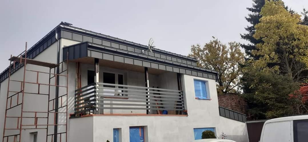 Dom w trakcie remontu z nowym, grafitowym dachem blaszanym, widoczne rusztowania i okna zabezpieczone folią.