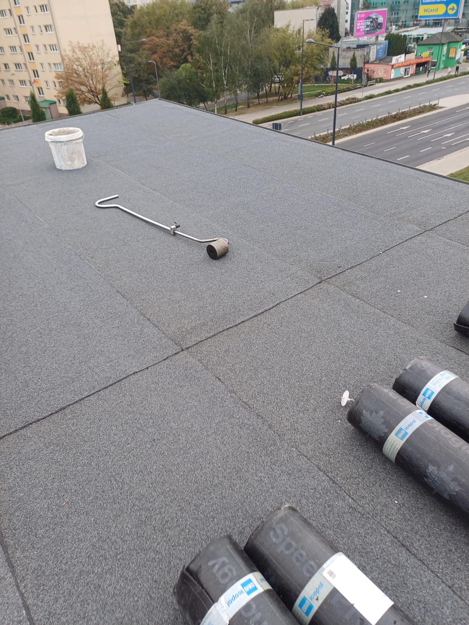 Szary, płaski dach pokryty papą termozgrzewalną, z widocznymi rolkami papy BMI Icopal Speedway, narzędziem dekarskim do zgrzewania oraz wiadrem w tle, na tle miejskiego krajobrazu.