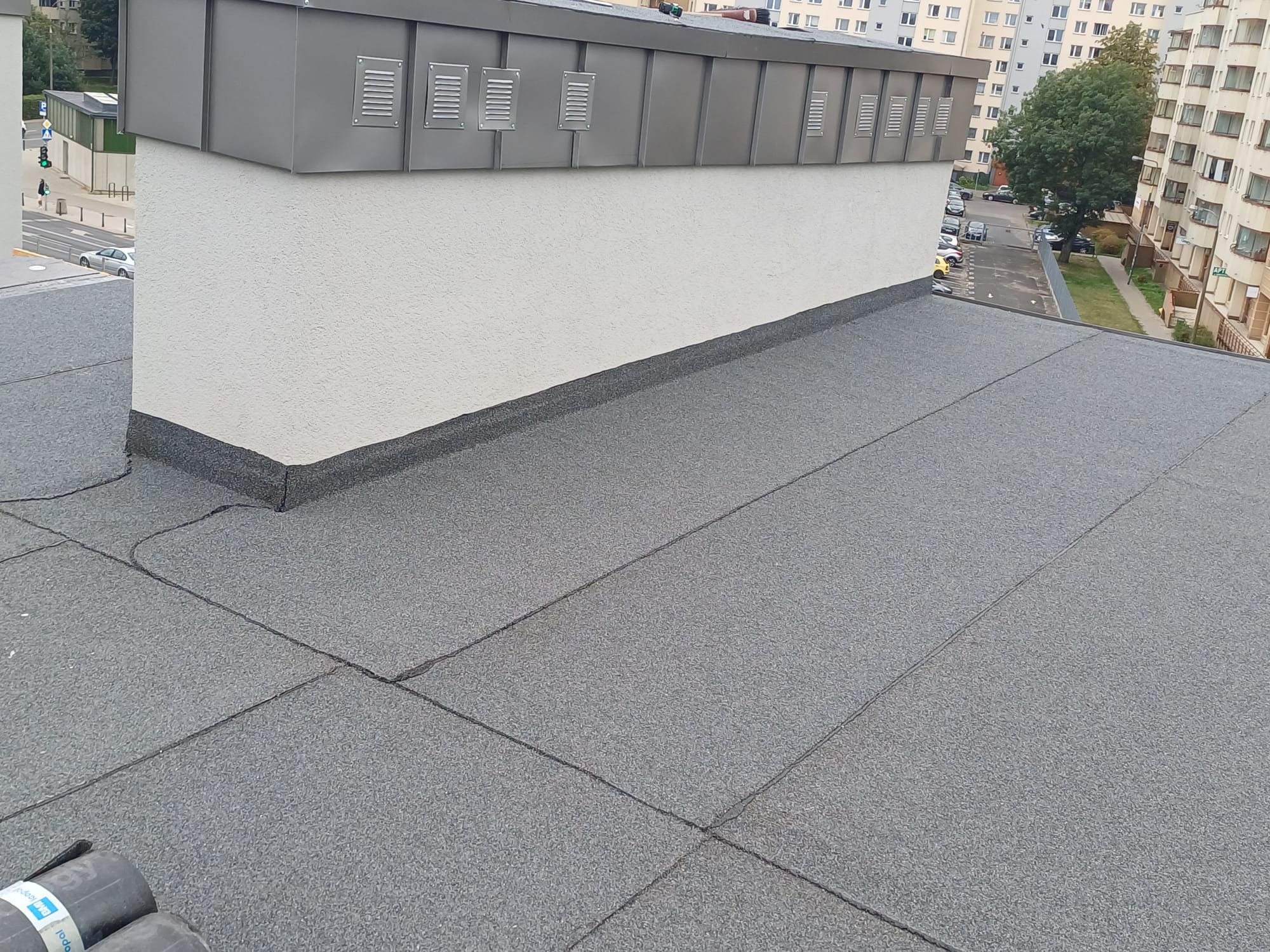 Szary dach pokryty papą termozgrzewalną na płaskim dachu budynku, widoczne kominy wentylacyjne i fragmenty otoczenia miejskiego w tle.