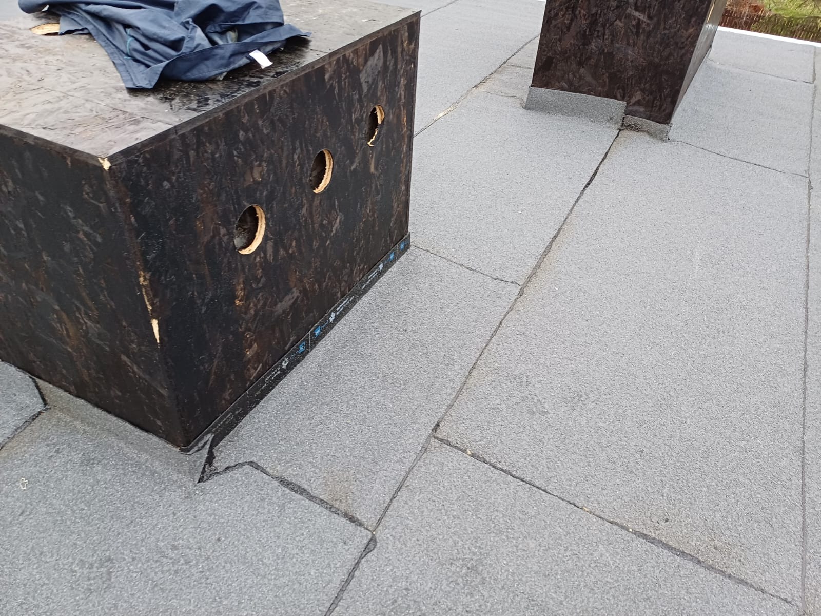 Szary dach pokryty papą z czarnym kominem z otworami wentylacyjnymi, widoczne łączenia papy.