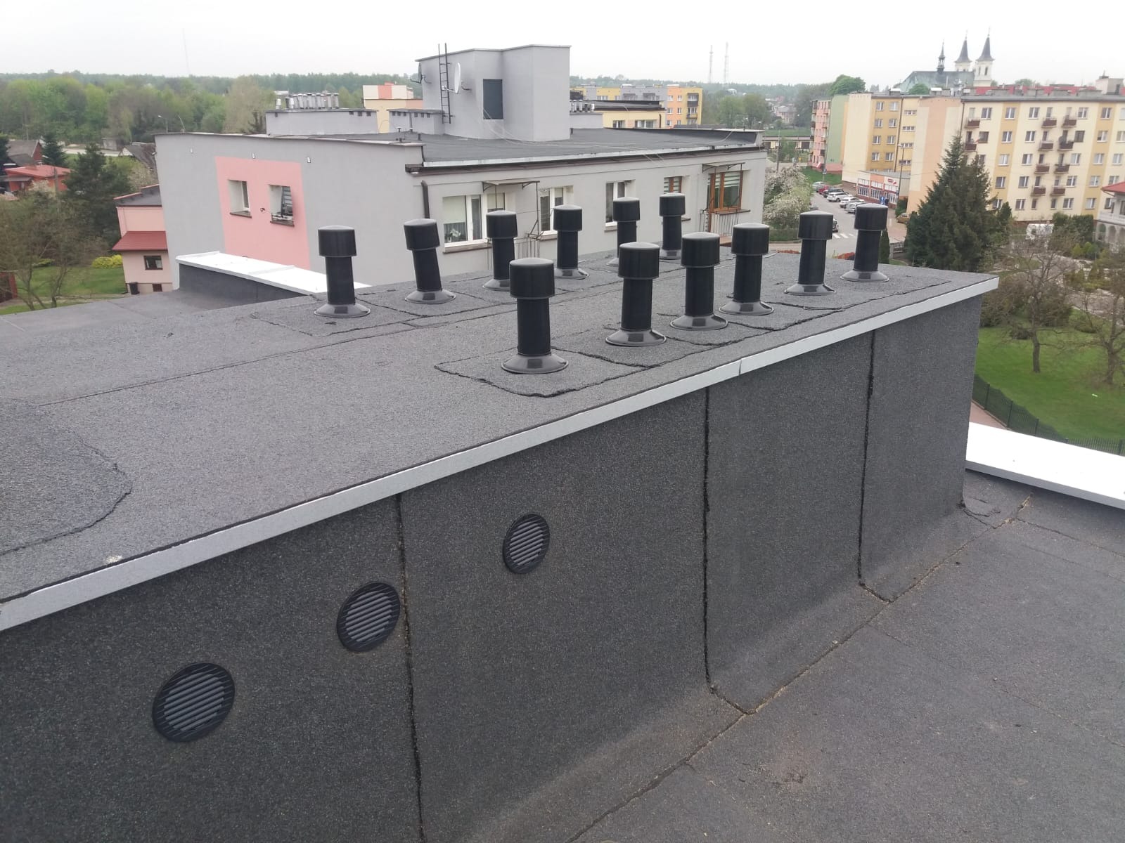 Płaski dach pokryty papą z licznymi czarnymi kominkami wentylacyjnymi i okrągłymi kratkami wentylacyjnymi na attyce, w tle zabudowa miejska.
