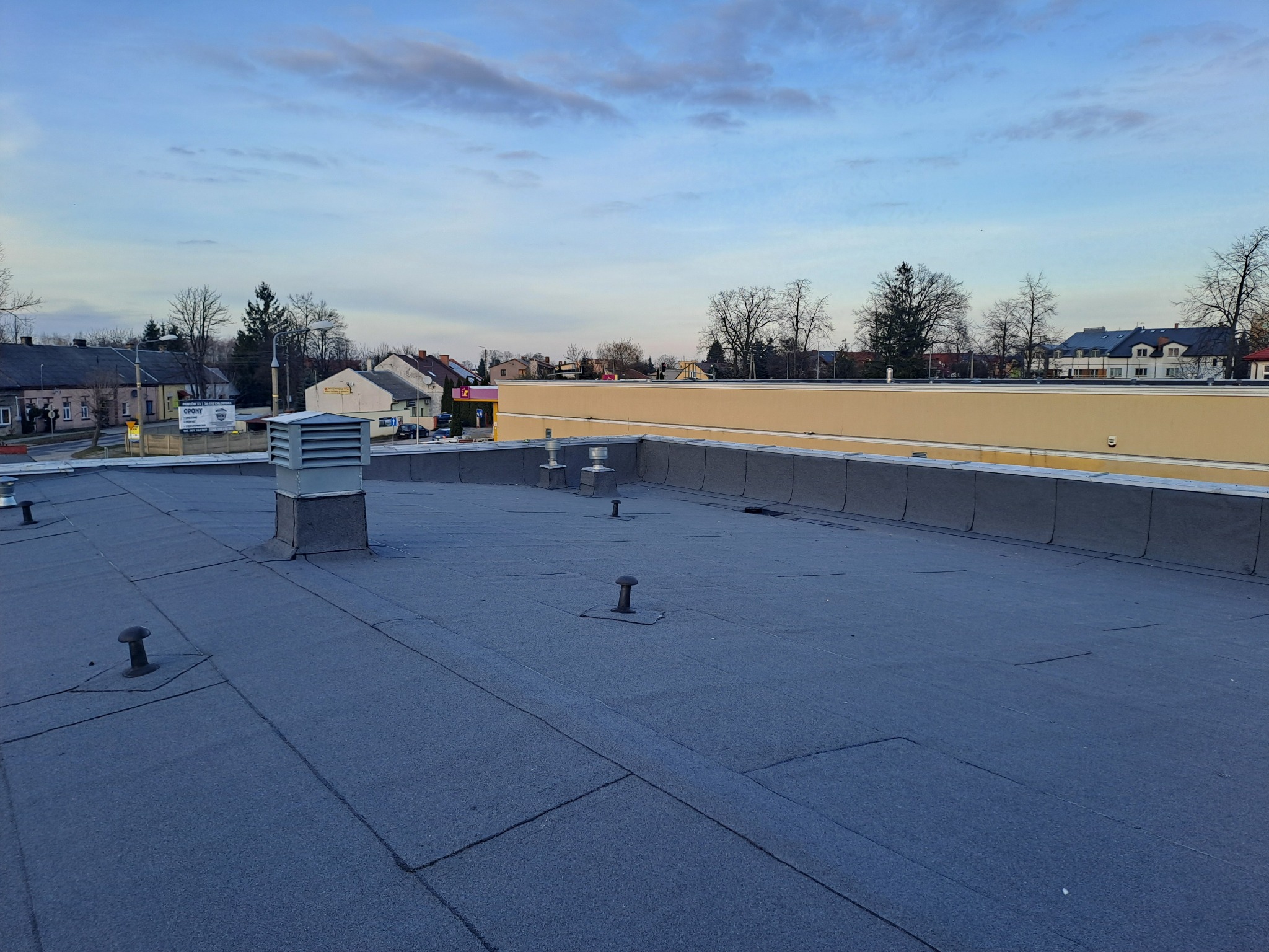 Płaski dach pokryty papą termozgrzewalną z widocznymi kominkami wentylacyjnymi i kominem, na tle zabudowań miejskich pod pochmurnym niebem.