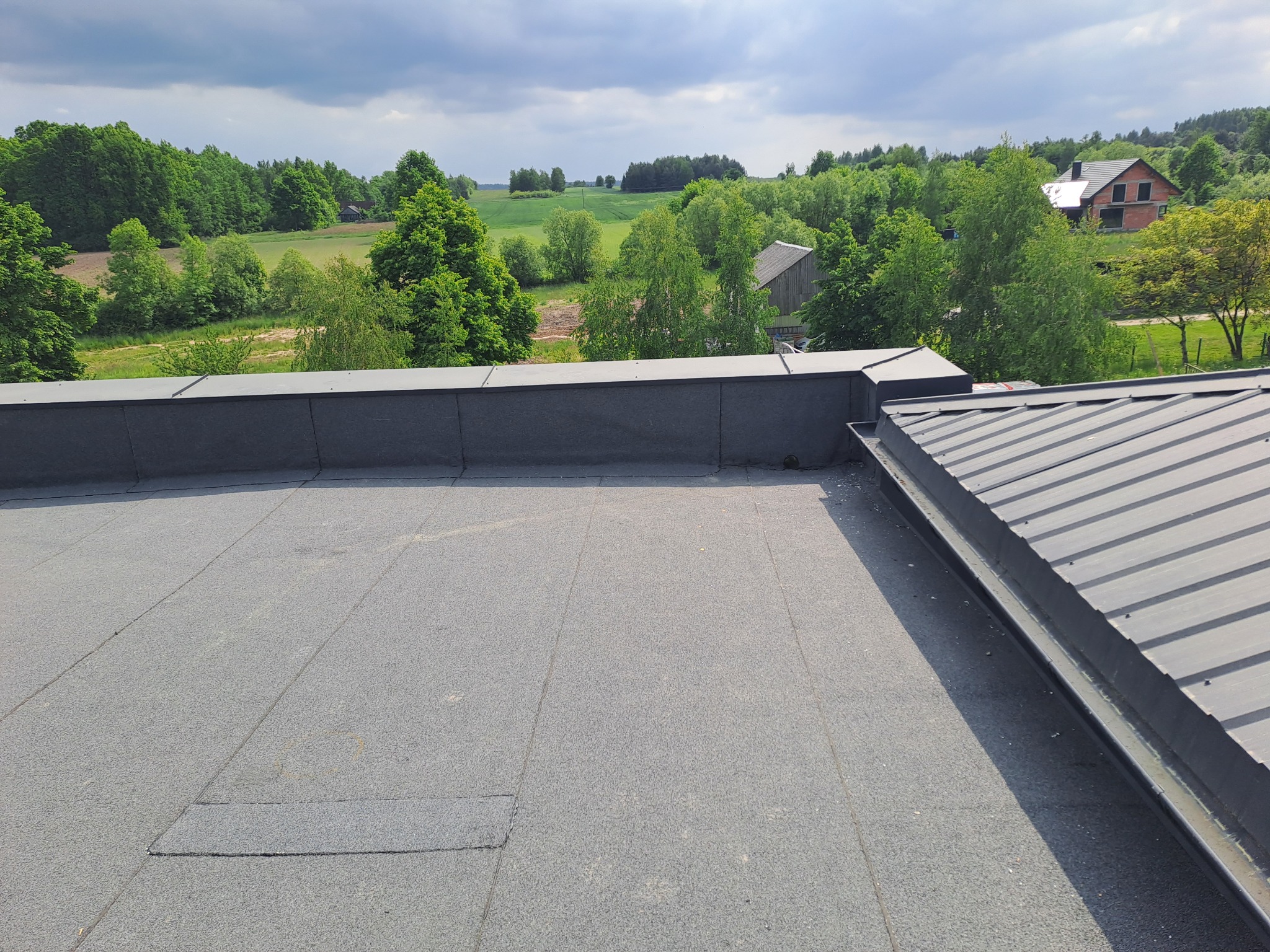 Widok z góry na płaski dach pokryty papą termozgrzewalną, z widocznymi łączeniami i elementami wykończeniowymi, w tle krajobraz wiejski z polami, lasami i budynkami.