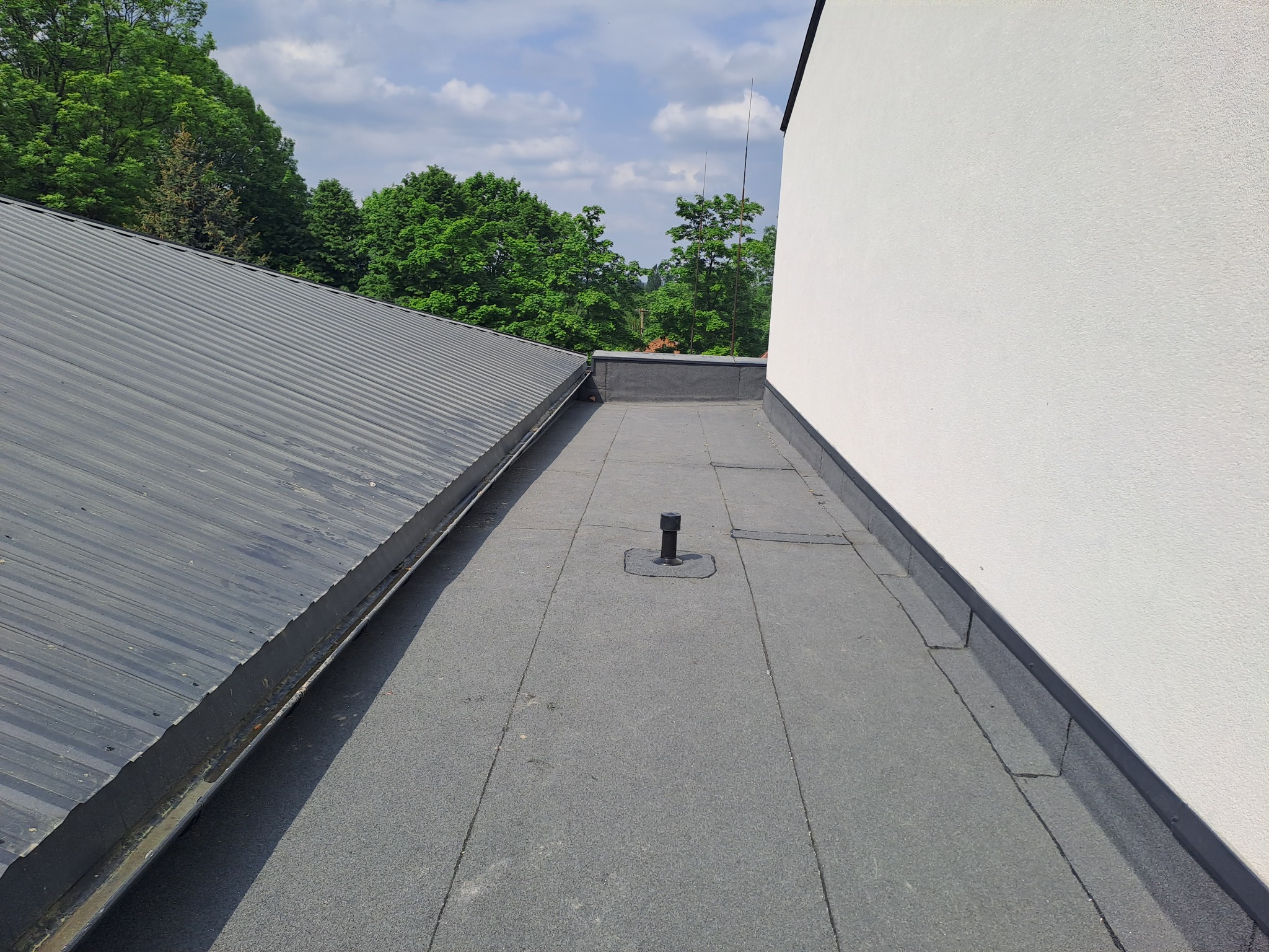 Widok z góry na płaski dach pokryty papą, z widocznym kominkiem wentylacyjnym i fragmentem dachu skośnego z blachy trapezowej oraz białą ścianą budynku w tle.