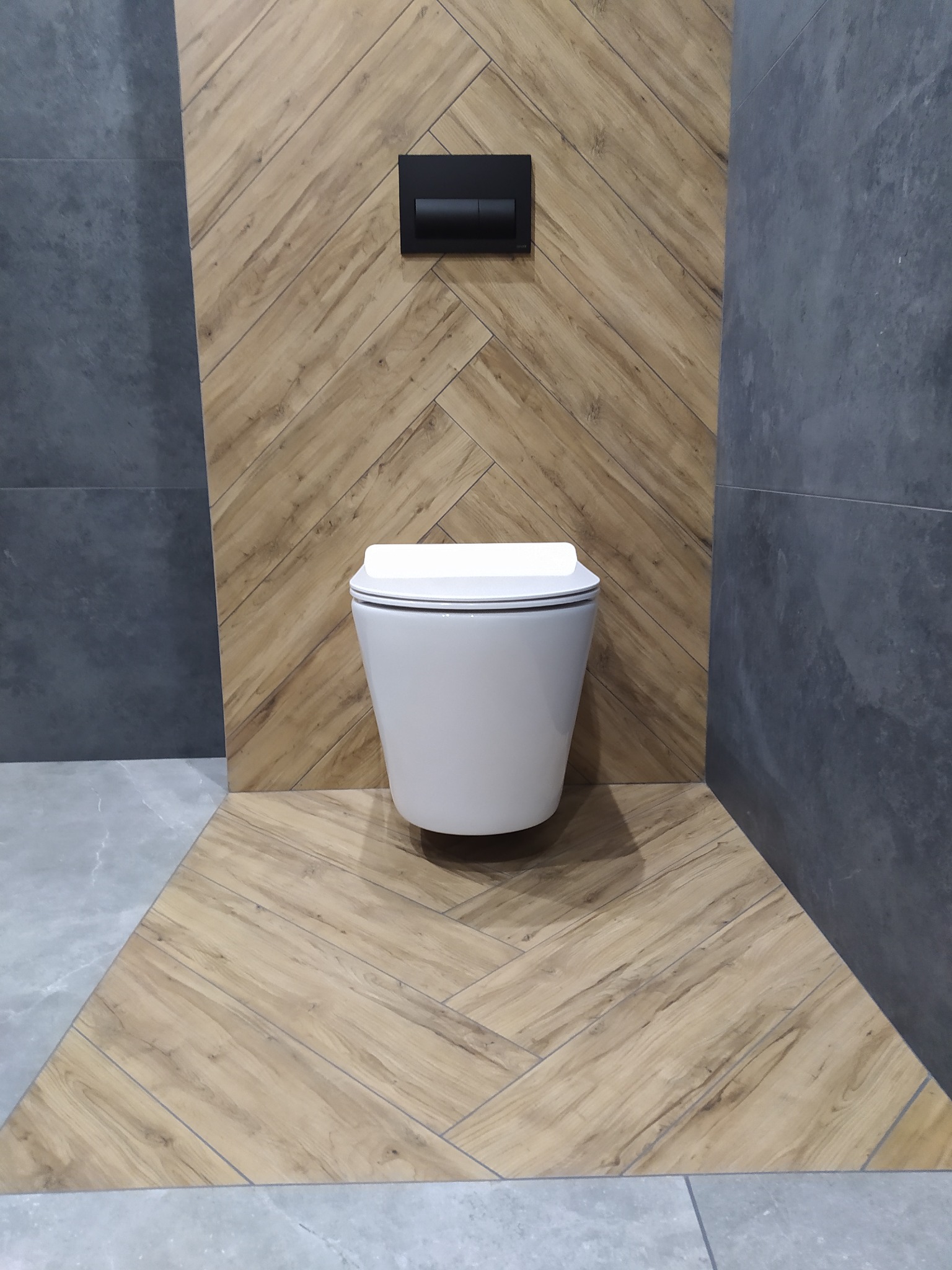 Wisząca toaleta z czarnym przyciskiem spłukującym na ścianie wyłożonej panelami drewnopodobnymi ułożonymi w jodełkę, obok ściany z szarych płytek.
