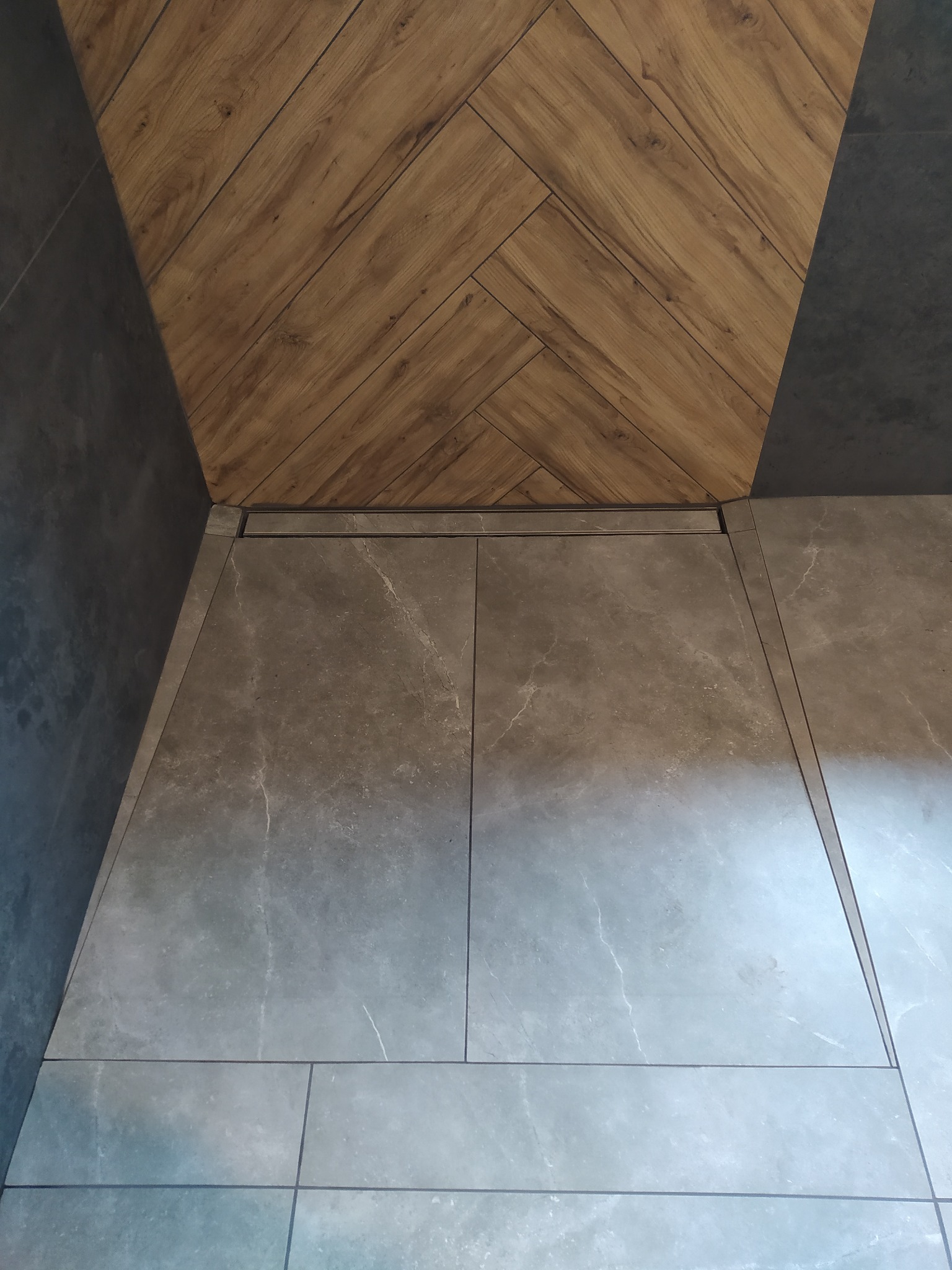 Szare płytki podłogowe z delikatnym wzorem marmuru, ułożone w prostokątnym formacie, stykają się z drewnopodobną okładziną ścienną ułożoną w jodełkę i ciemnoszarą okładziną ścienną, tworząc...