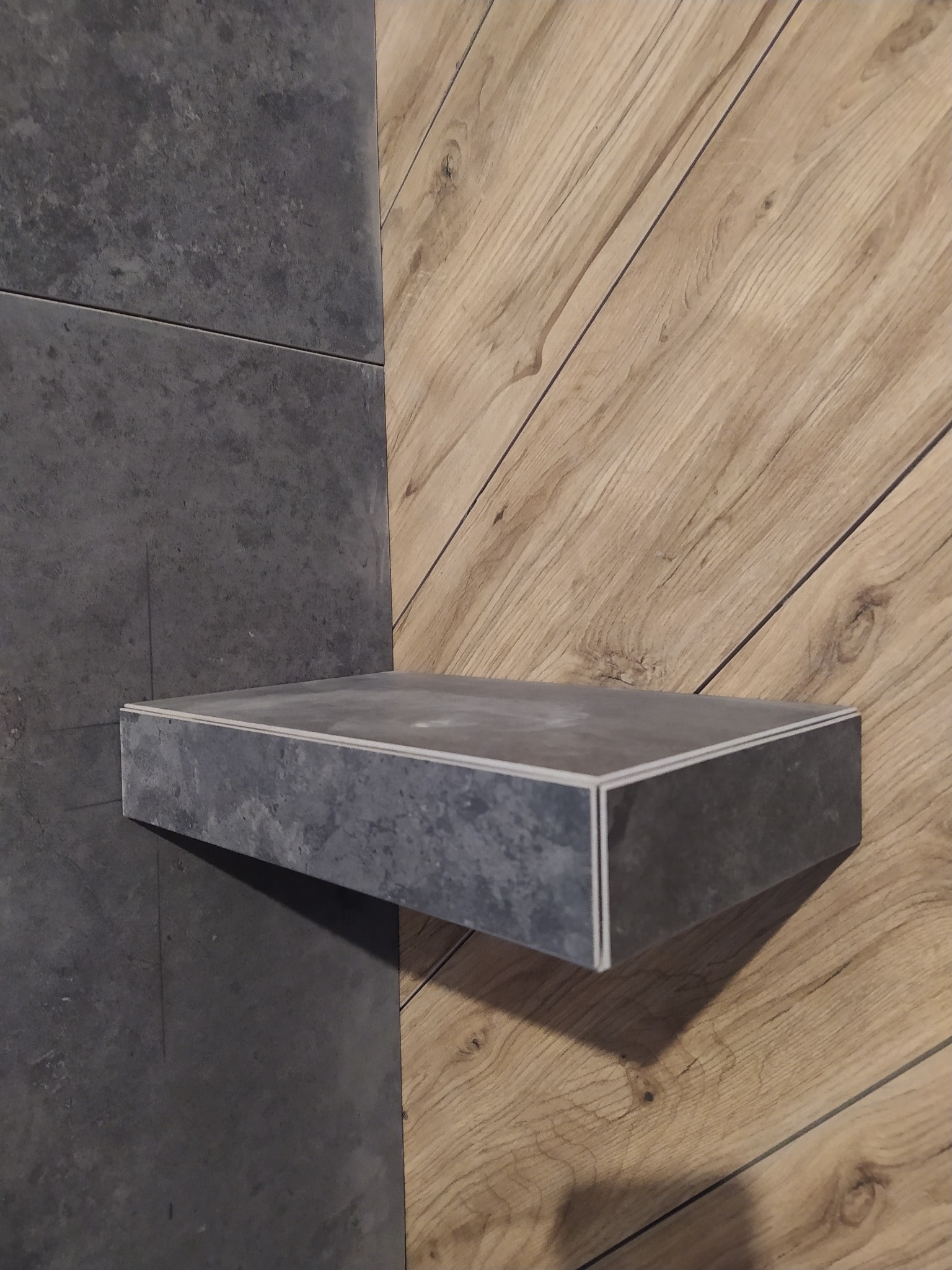 Półka z ciemnoszarej płyty ceramicznej z jasną krawędzią, zamontowana na ścianie z ciemnoszarych płytek, z widocznym fragmentem ściany wyłożonej panelami imitującymi drewno.