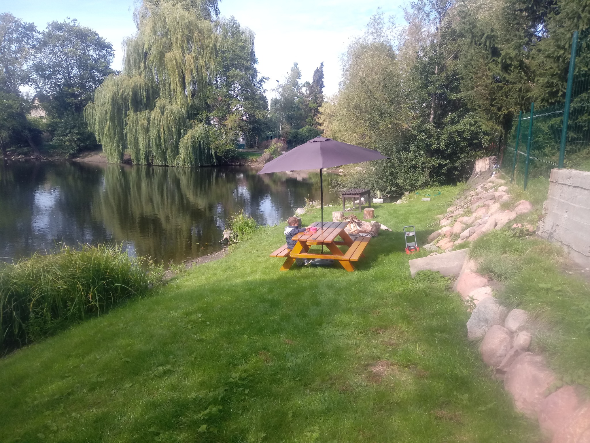 Drewniany stół piknikowy z parasolem nad brzegiem stawu, otoczony zielenią i drzewami.