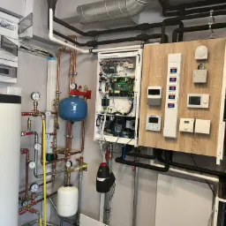 Złożona instalacja grzewcza z kotłem, zbiornikiem buforowym, naczyniami wyr&oacute;wnawczymi i panelami sterowania zamontowanymi na ścianie.