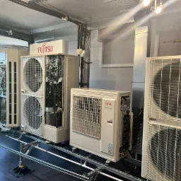 Rząd białych jednostek zewnętrznych klimatyzacji marki Fujitsu, niektóre z otwartymi obudowami ukazującymi wewnętrzne komponenty, ustawionych na metalowej konstrukcji w pomieszczeniu z oknem.