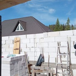 Budowlani - Wznoszenie ściany z białych bloczków na budowie domu jednorodzinnego; widoczni dwaj pracownicy, rusztowanie, drabina, zapas materiału i fragment dachu z oknem.