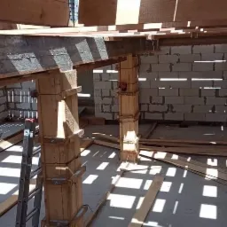 Wnętrze domu w trakcie budowy, widoczne ściany z bloczków, drewniane podpory pod stropem, rozsypane deski i drabina oparta o ścianę.