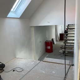 Budowlani - Wnętrze domu w trakcie prac wykończeniowych: surowe ściany, okno dachowe, tymczasowe schody, czerwone beczki i narzędzia na podłodze.