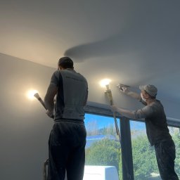 Budowlani - Dwóch mężczyzn w roboczych ubraniach wyrównuje gładź na suficie przy użyciu szpachli i lampy halogenowej, widok od dołu, okno z widokiem na zewnątrz w tle.
