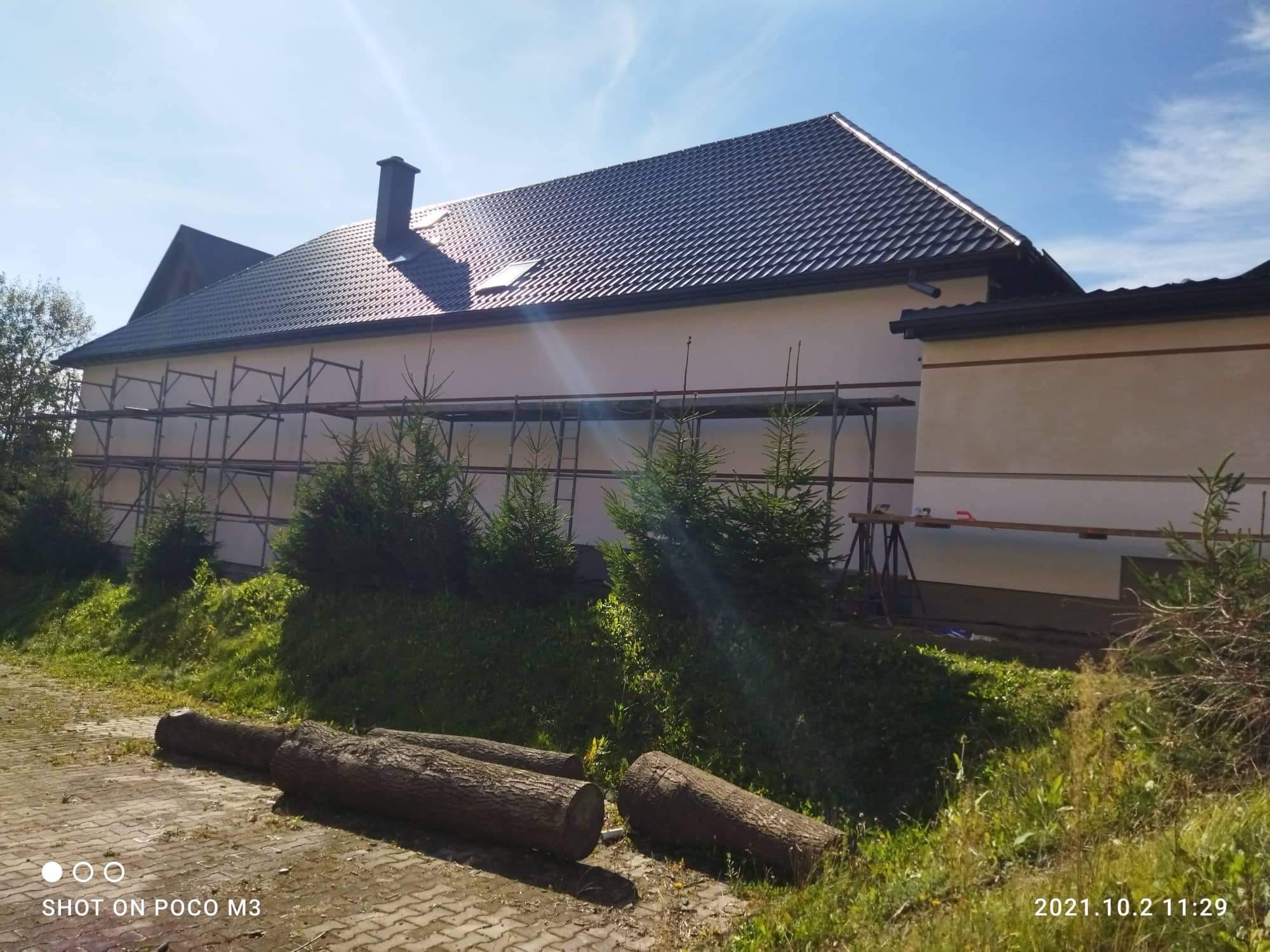 Dom jednorodzinny w trakcie prac elewacyjnych, widoczne rusztowania, świeżo otynkowana fasada w jasnym odcieniu oraz ciemna dachówka, na pierwszym planie trzy kłody drewna.