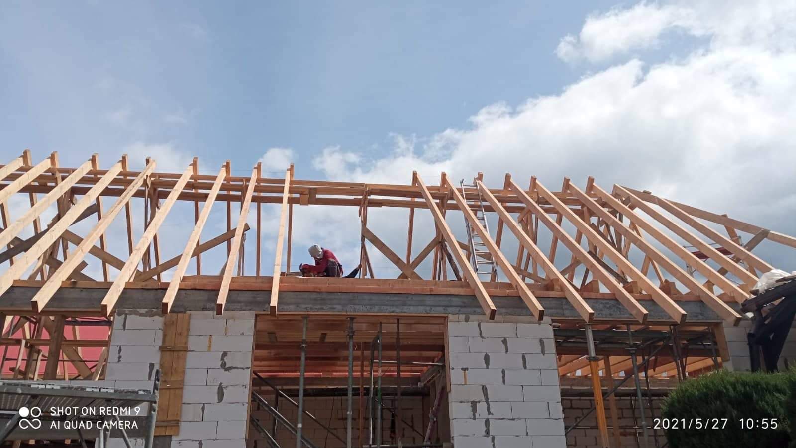 Wznoszenie drewnianej konstrukcji dachu na murowanym domu z betonu komórkowego, widoczny pracownik w czerwonej koszuli i kasku, drabina oparta o konstrukcję, błękitne niebo w tle.