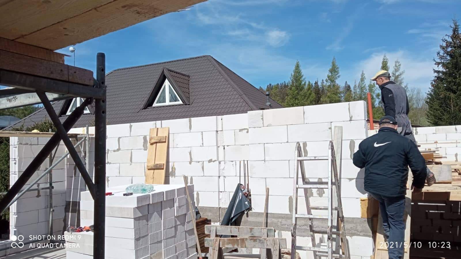 Wznoszenie ściany z białych bloczków na budowie domu jednorodzinnego; widoczni dwaj pracownicy, rusztowanie, drabina, zapas materiału i fragment dachu z oknem.