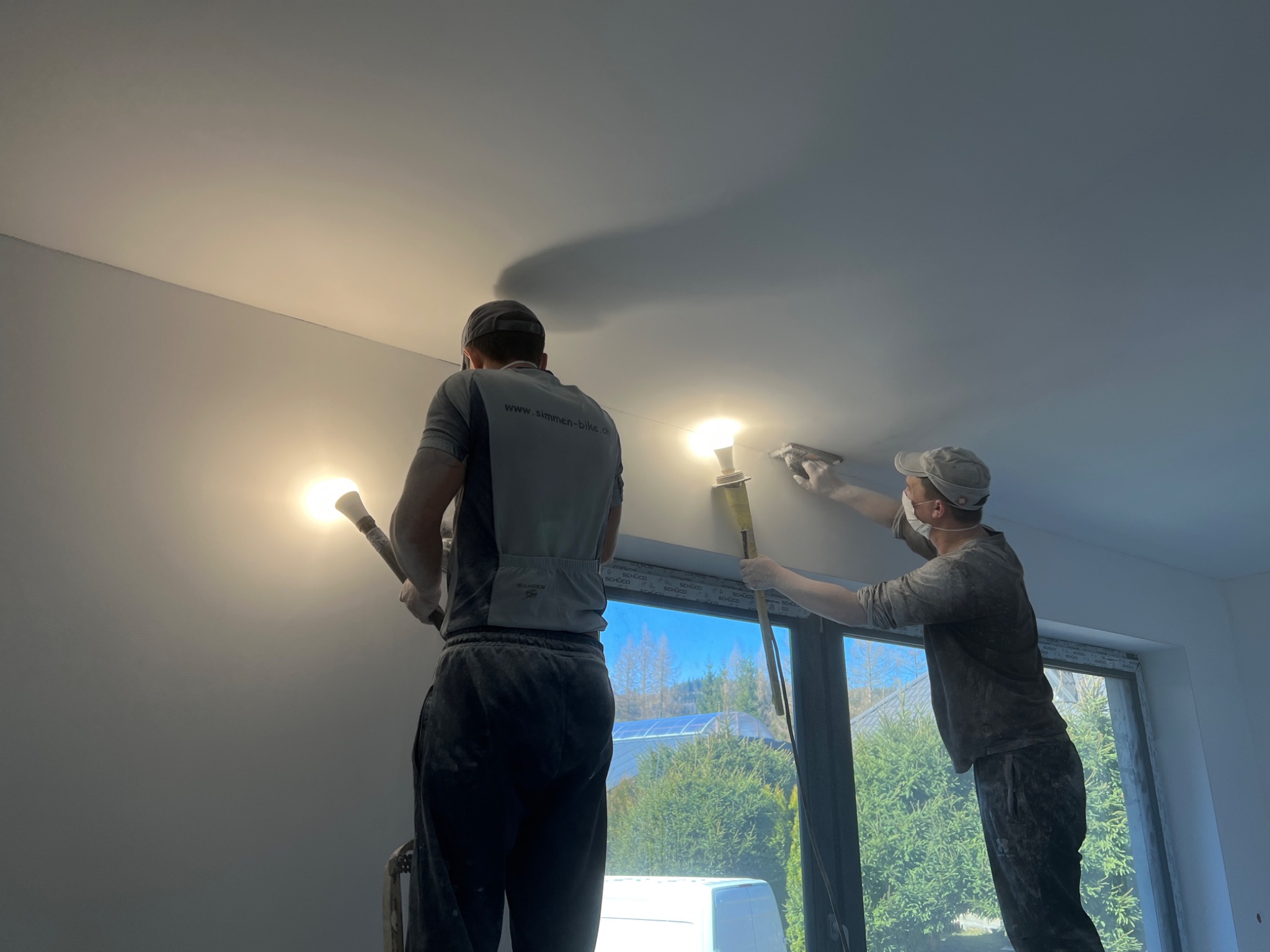 Dwóch mężczyzn w roboczych ubraniach wyrównuje gładź na suficie przy użyciu szpachli i lampy halogenowej, widok od dołu, okno z widokiem na zewnątrz w tle.