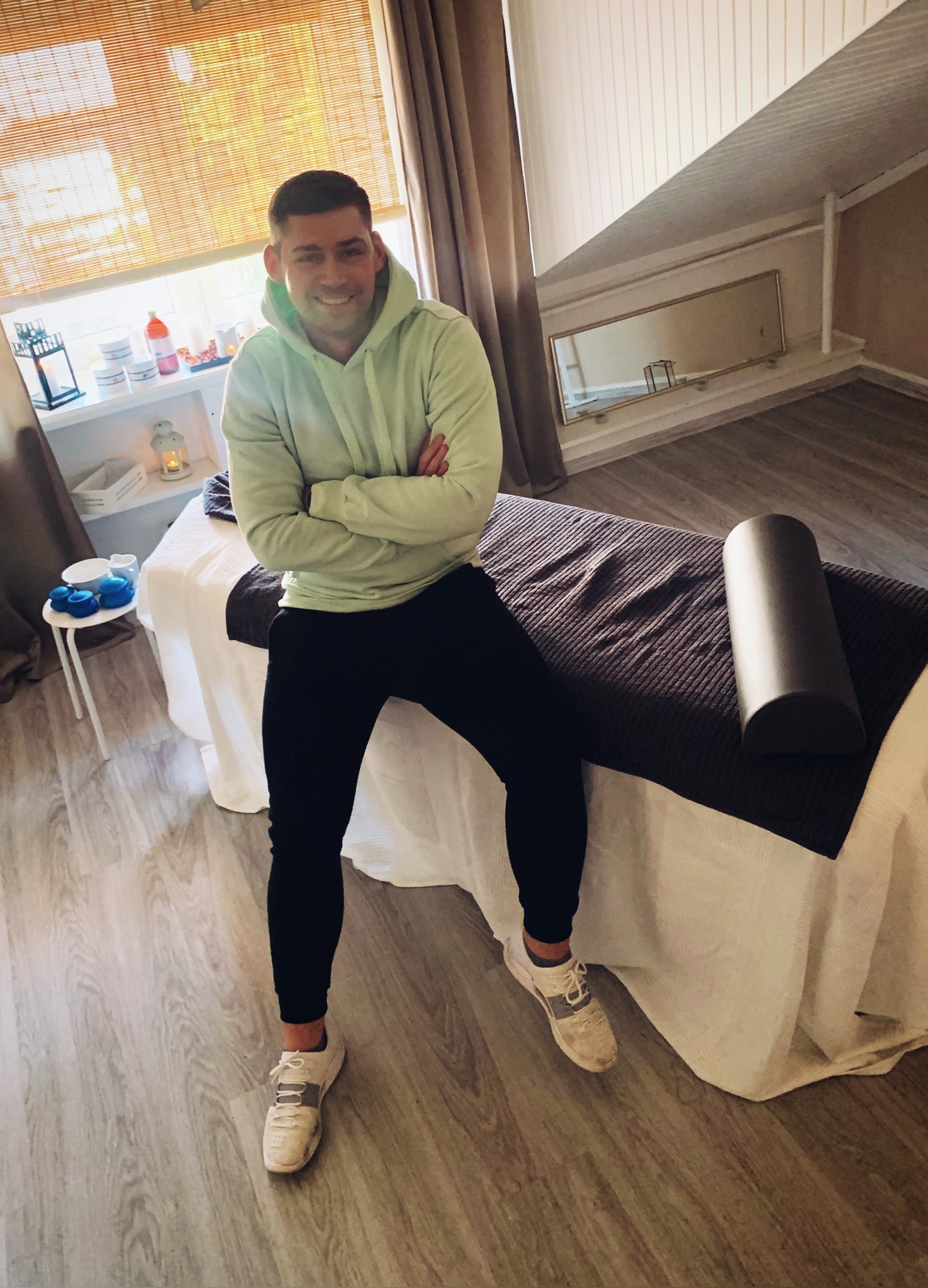 Uśmiechnięty mężczyzna w bluzie z kapturem siedzi na stole do zabiegów z zagłówkiem, w tle okno z żaluzjami i kosmetyki na półce.