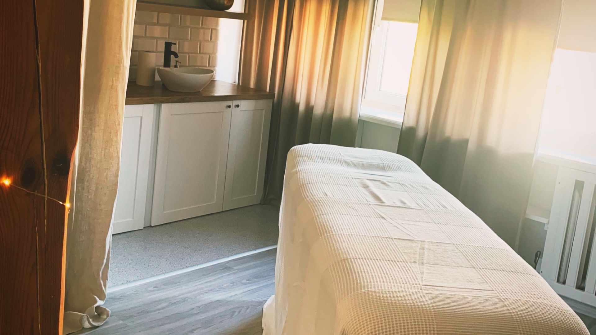 Jasne wnętrze gabinetu z łóżkiem do zabiegów przykrytym białą kołdrą, umywalką na szafce z białymi frontami i zasłoniętym oknem, oświetlone ciepłym światłem słonecznym.