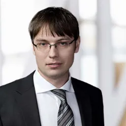 Portret adwokata w okularach, w czarnym garniturze i krawacie w szaro-białe paski, na rozmytym tle biura.